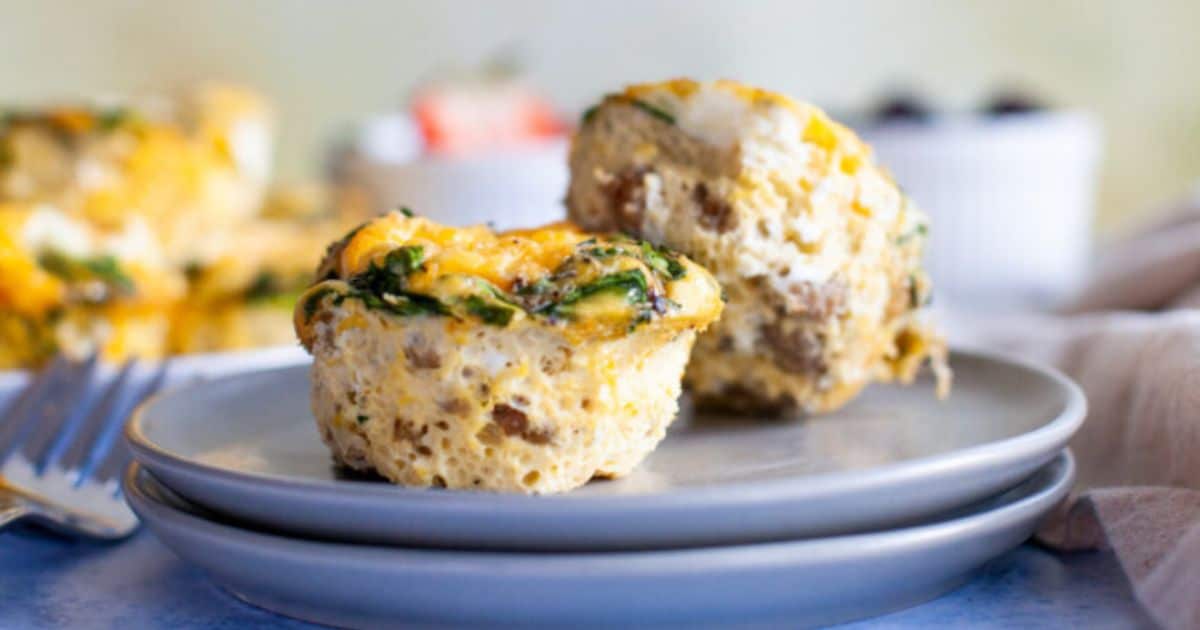 Two egg muffin cups with visible spinach and sausage sit on stacked plates; one muffin is cut open to show the inside. A fork and blurred food items are in the background.