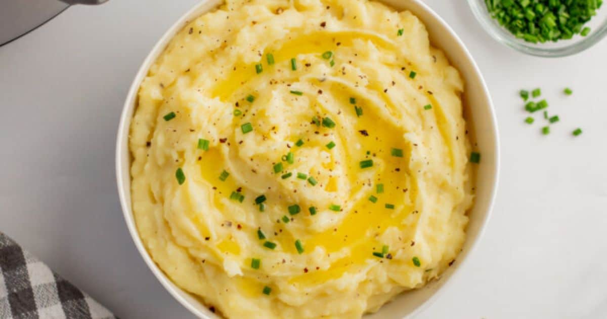 A bowl of mashed potatoes topped with melted butter, chopped chives, and ground black pepper, placed on a white surface.