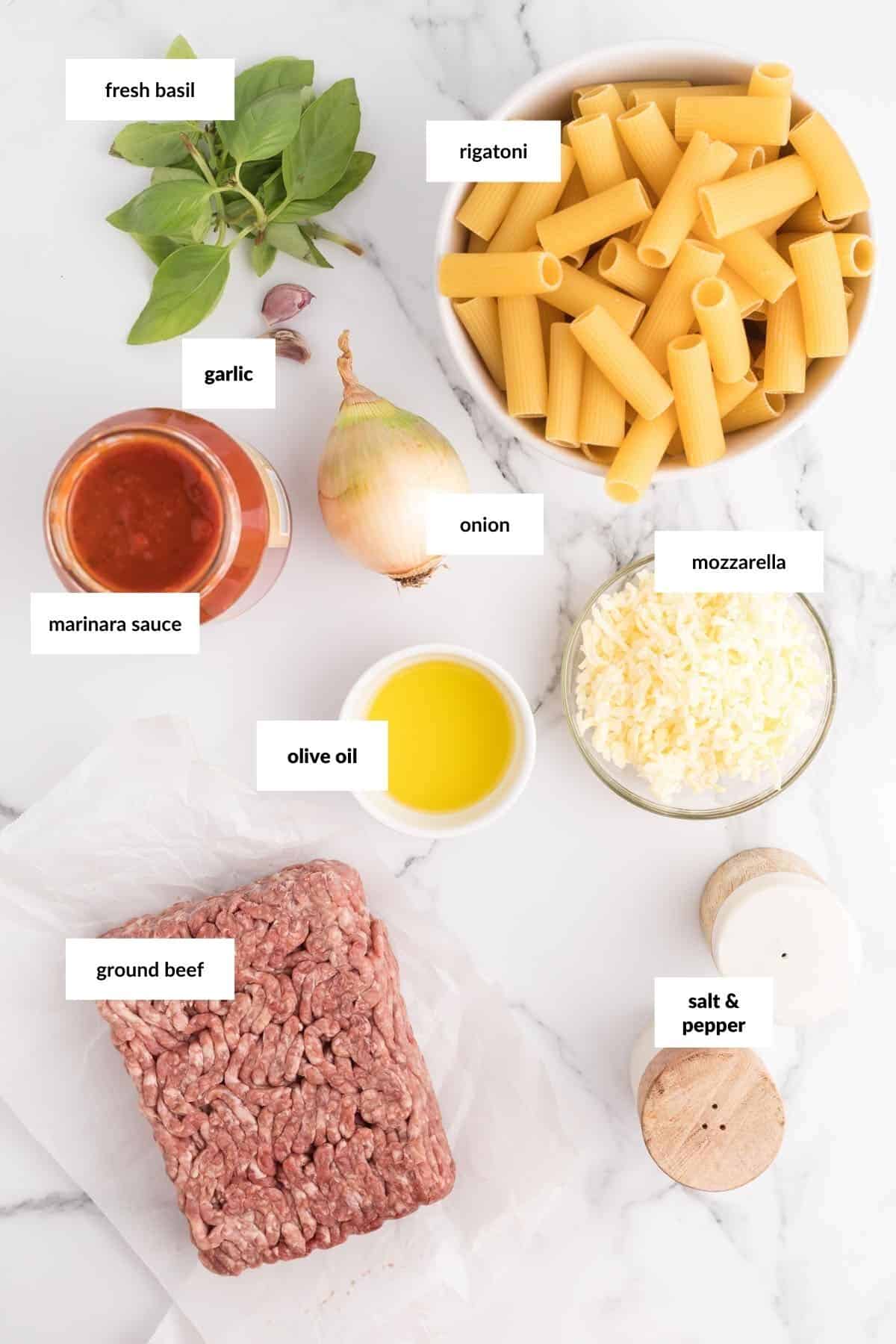 Ingredients for a pasta dish: uncooked rigatoni, ground beef, mozzarella cheese, onion, garlic, fresh basil, marinara sauce, olive oil, salt, and pepper arranged on a white surface.