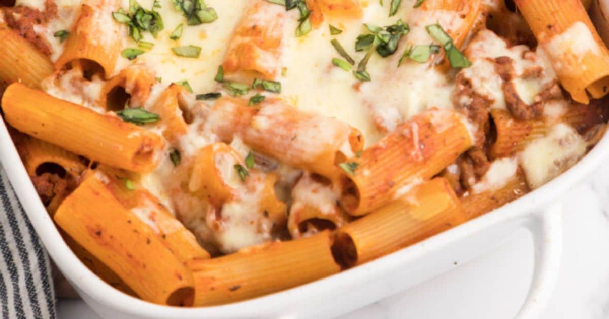 Close-up of baked rigatoni pasta with melted cheese, tomato sauce, ground meat, and chopped herbs in a white casserole dish.