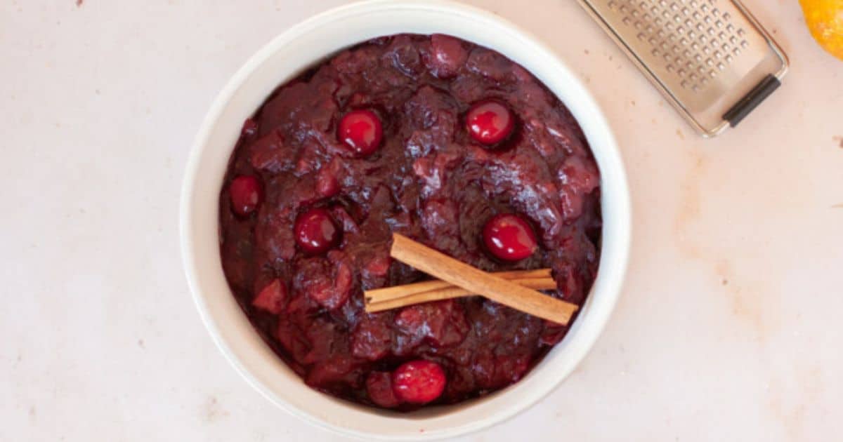 A white bowl filled with homemade cranberry sauce, topped with whole cranberries and two cinnamon sticks, sits on a light countertop next to a metal grater.