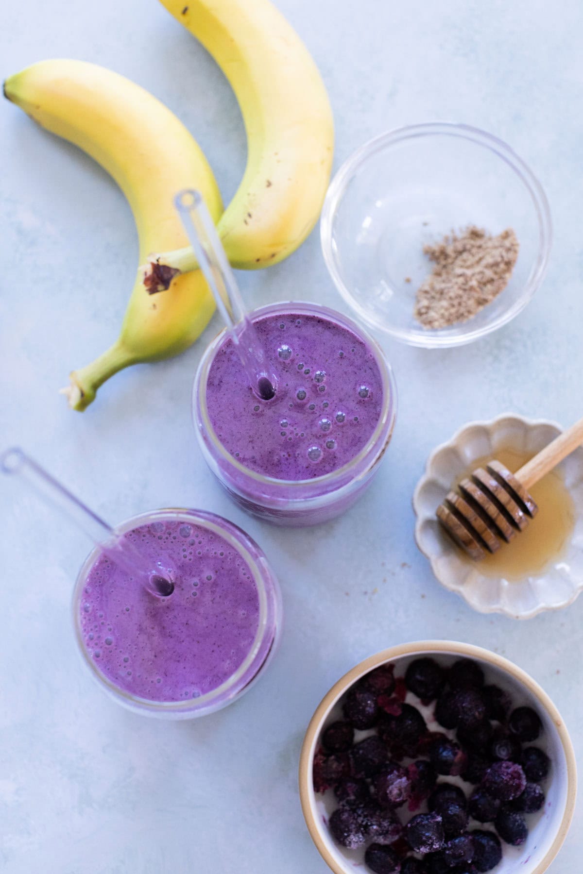 Two glasses of a banana blueberry smoothie with straws, surrounded by two bananas, a bowl of mixed berries, a small dish of honey with a dipper, and a bowl of ground flaxseed.