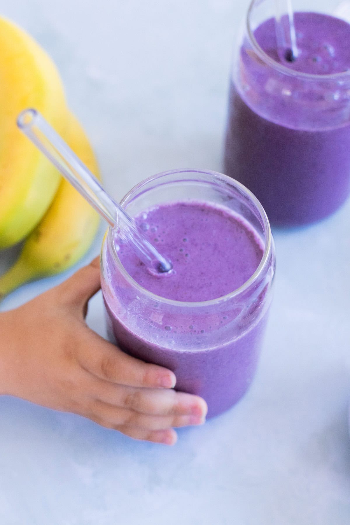 A child&rsquo;s hand holding a glass of banana blueberry smoothie with a clear straw, next to another smoothie and bananas on a light surface.