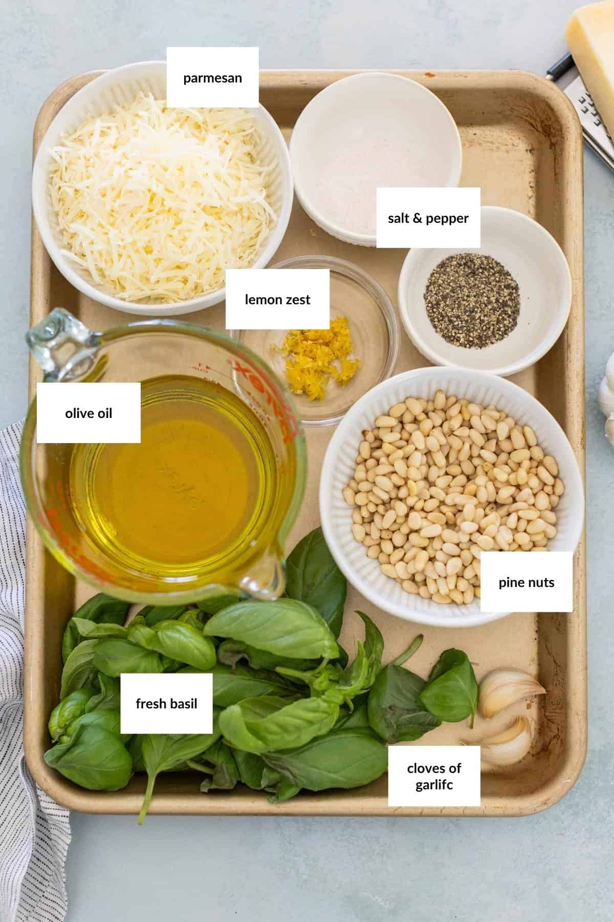 A tray with bowls containing parmesan, salt & pepper, lemon zest, olive oil, pine nuts, fresh basil, and cloves of garlic, each labeled with text.