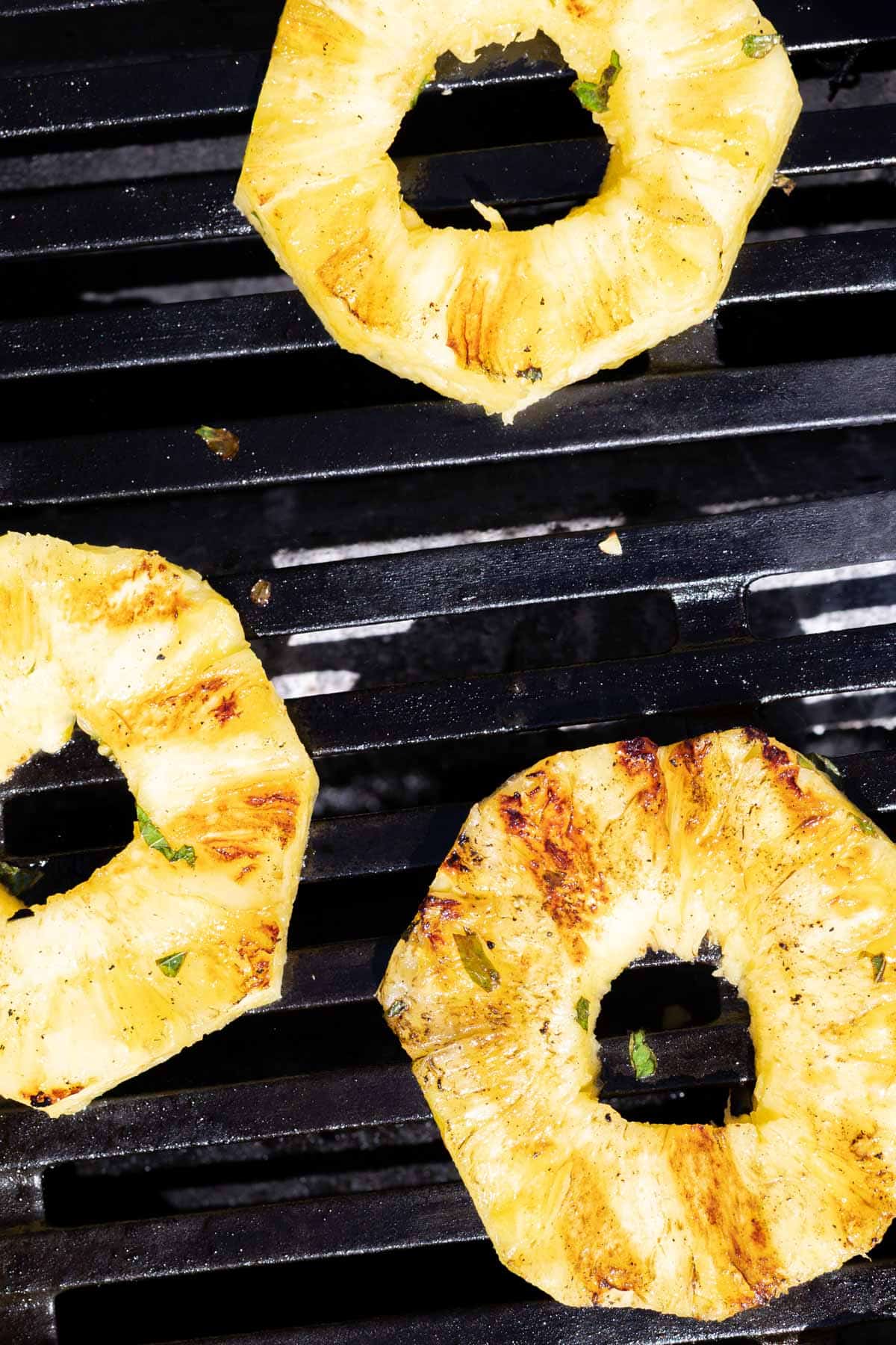 Three pineapple rings with grill marks are cooking on a black barbecue grill.
