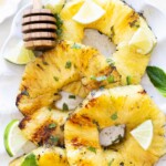 Grilled pineapple slices garnished with lime wedges and mint leaves, served on a white plate with a honey dipper.