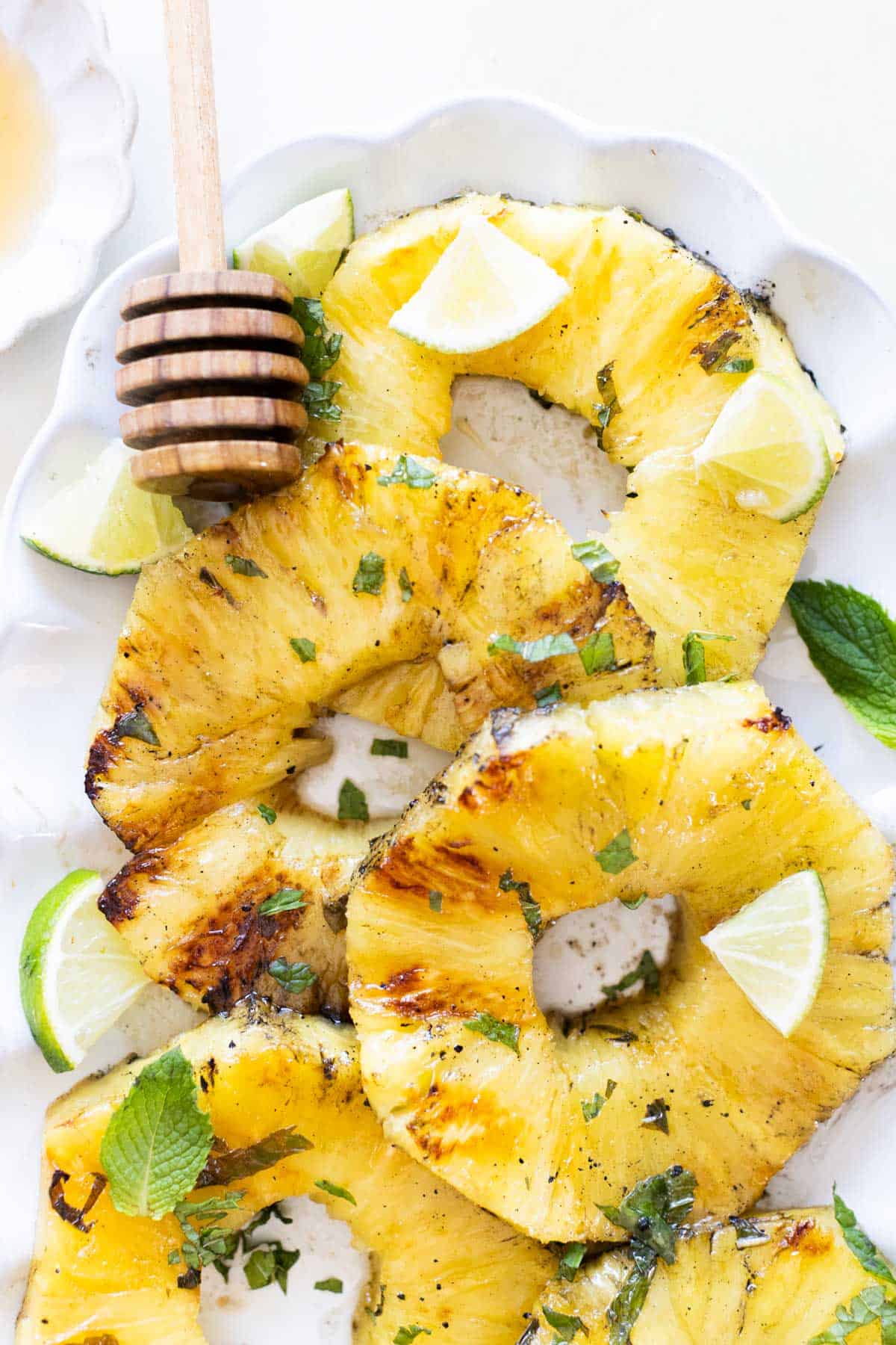 Grilled pineapple slices garnished with lime wedges and mint leaves, served on a white plate with a honey dipper.