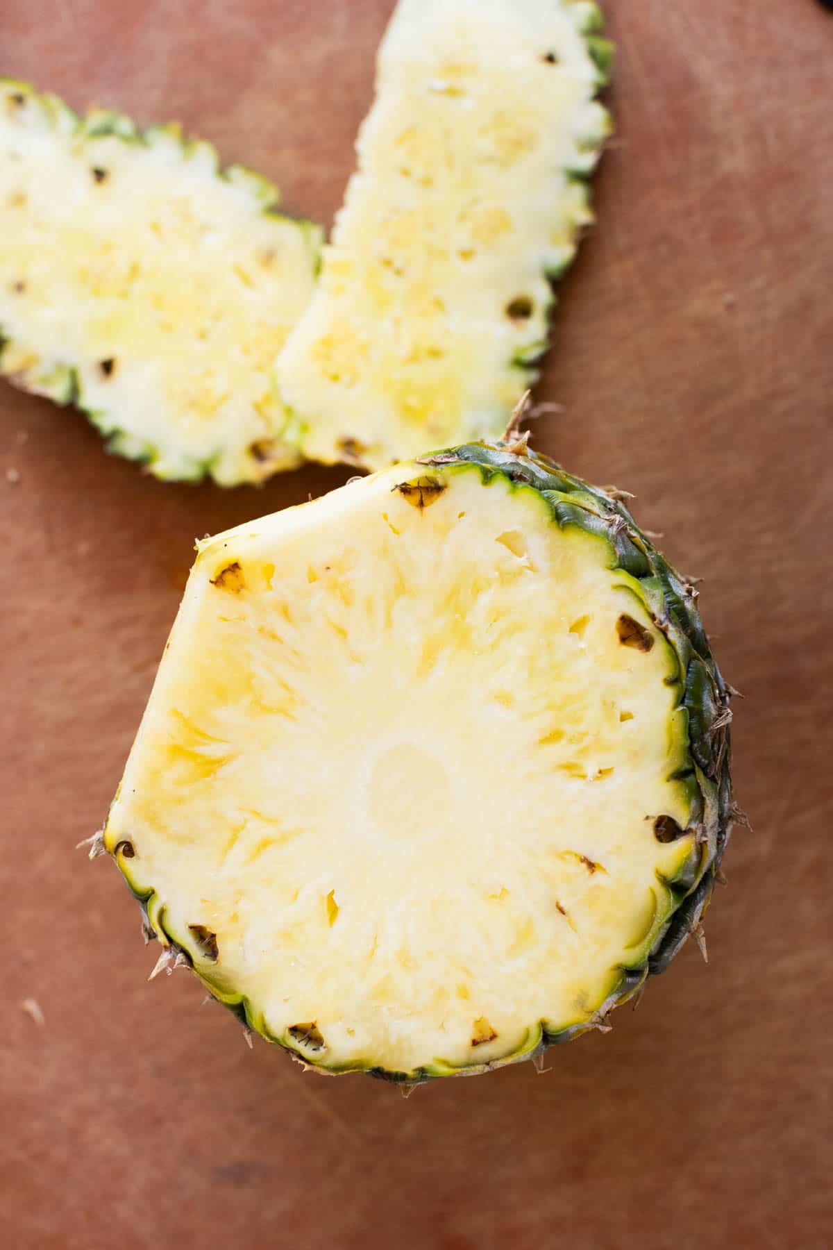 A sliced pineapple with the top cut off and two thin slices of pineapple rind nearby on a wooden surface.