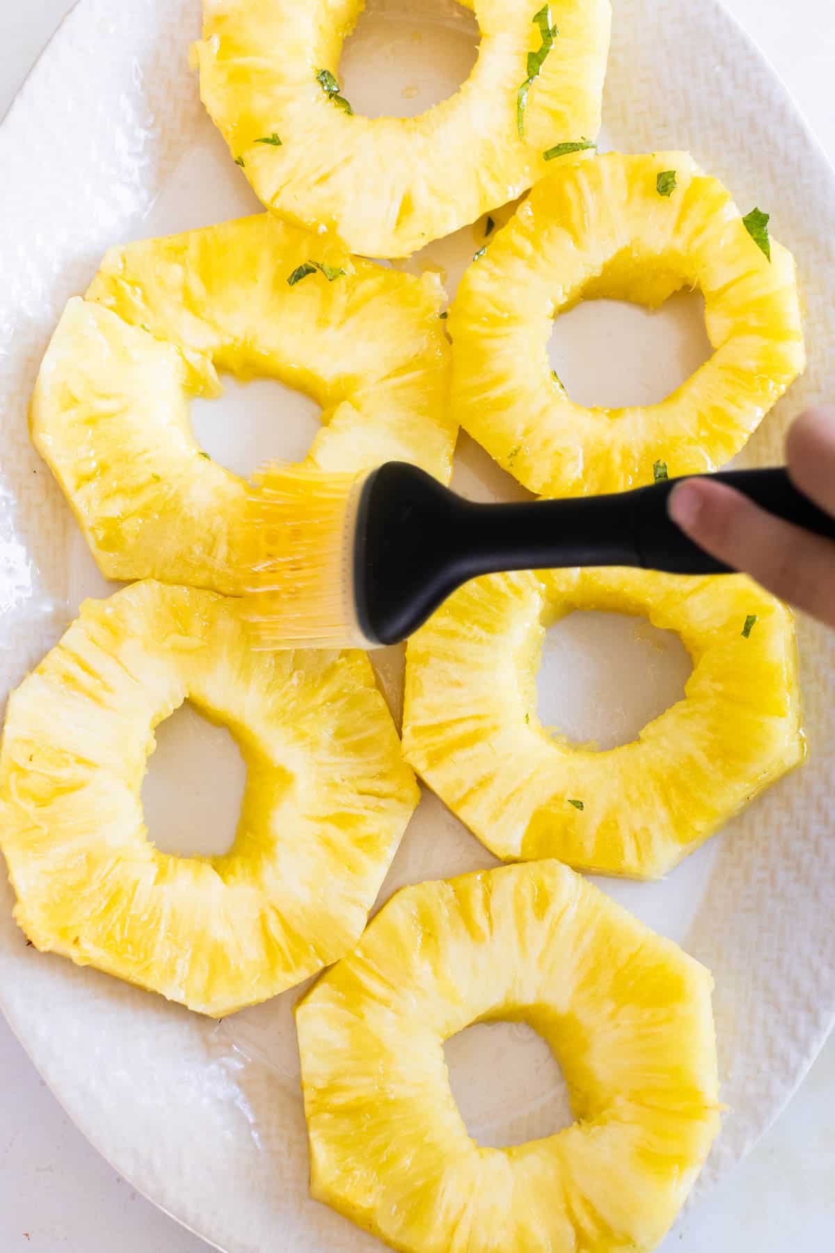 A hand uses a brush to coat pineapple rings with seasoned melted butter arranged on a white plate.
