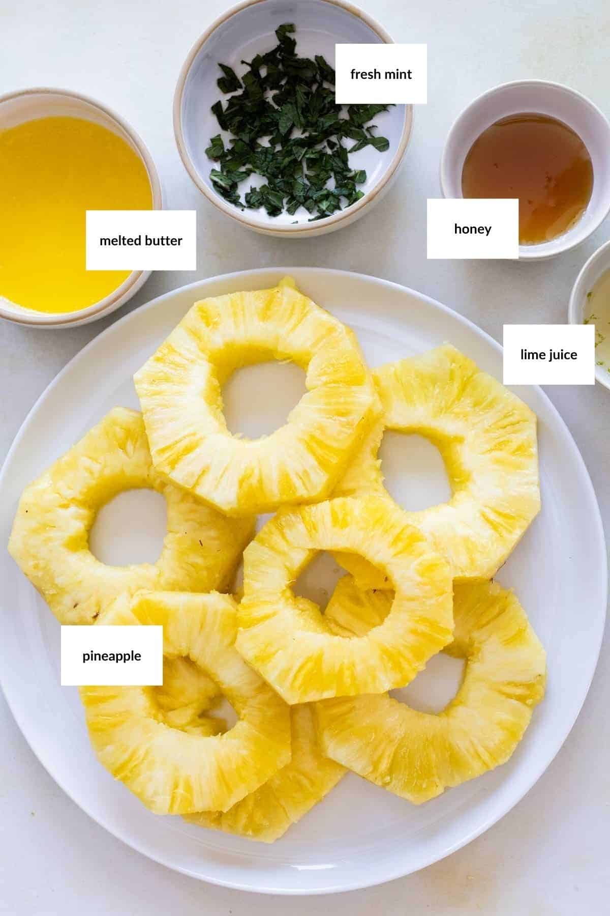 A white plate with grilled pineapple rings, surrounded by small bowls containing melted butter, chopped fresh mint, honey, and lime juice. Each ingredient is labeled.