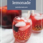 A glass of blackberry lemonade with ice and a straw, garnished with fresh blackberries and peach slices, sits on a light surface next to a pitcher and another glass.