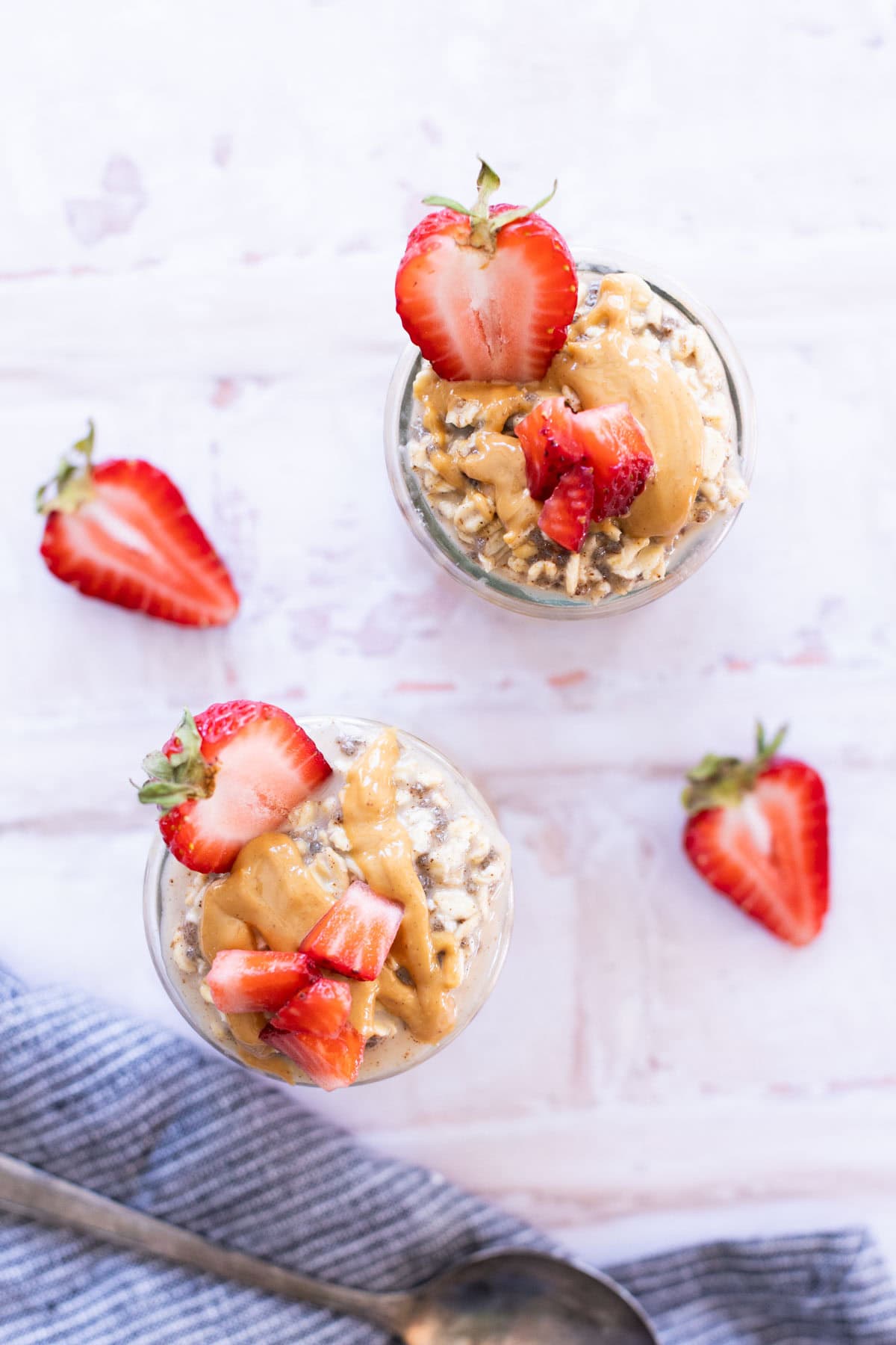 Two glass jars filled with overnight oats, topped with peanut butter and sliced strawberries, are placed on a light surface with extra strawberries nearby.