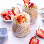 Two glass jars filled with overnight oats, topped with peanut butter and chopped strawberries, sit on a light surface with fresh strawberries nearby.