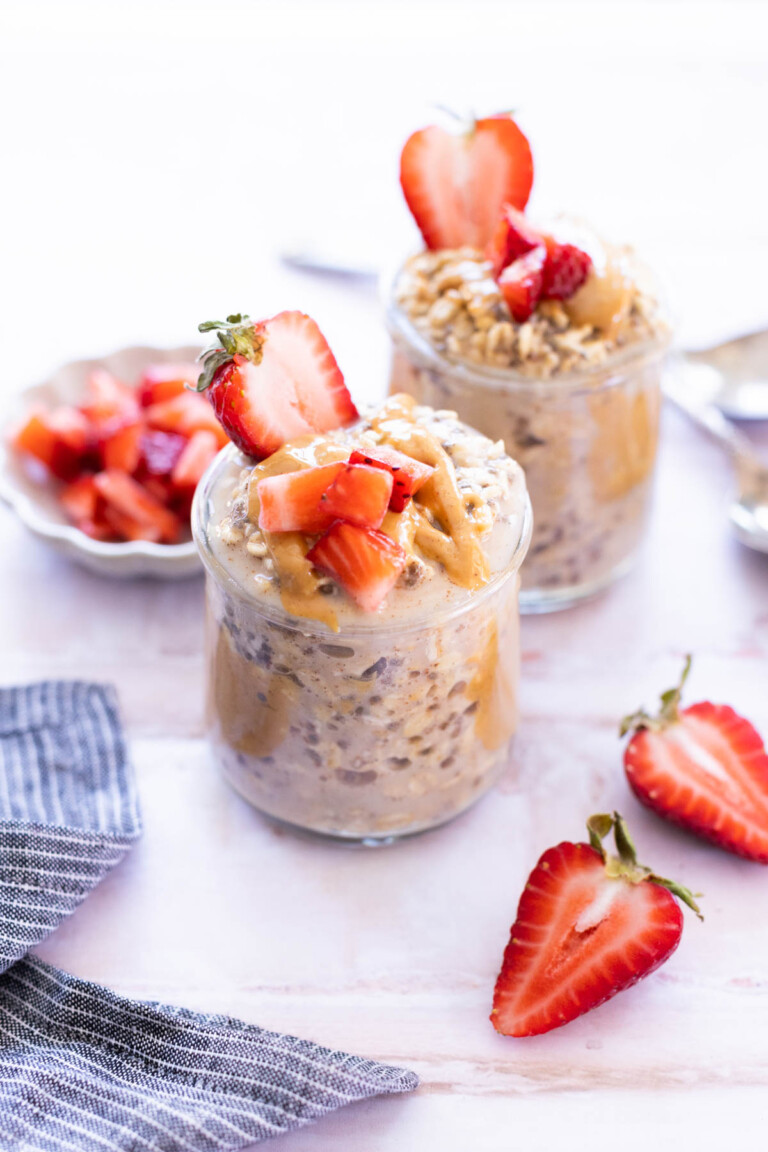 Two glass jars filled with overnight oats, topped with peanut butter and chopped strawberries, sit on a light surface with fresh strawberries nearby.