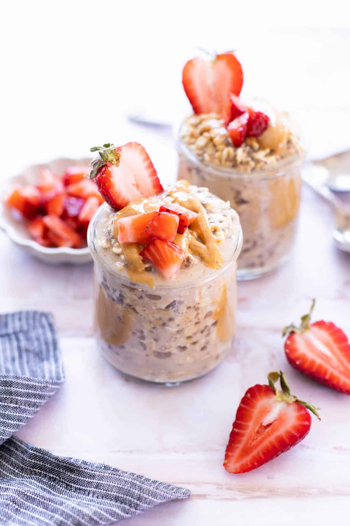 Two glass jars filled with overnight oats, topped with peanut butter and chopped strawberries, sit on a light surface with fresh strawberries nearby.