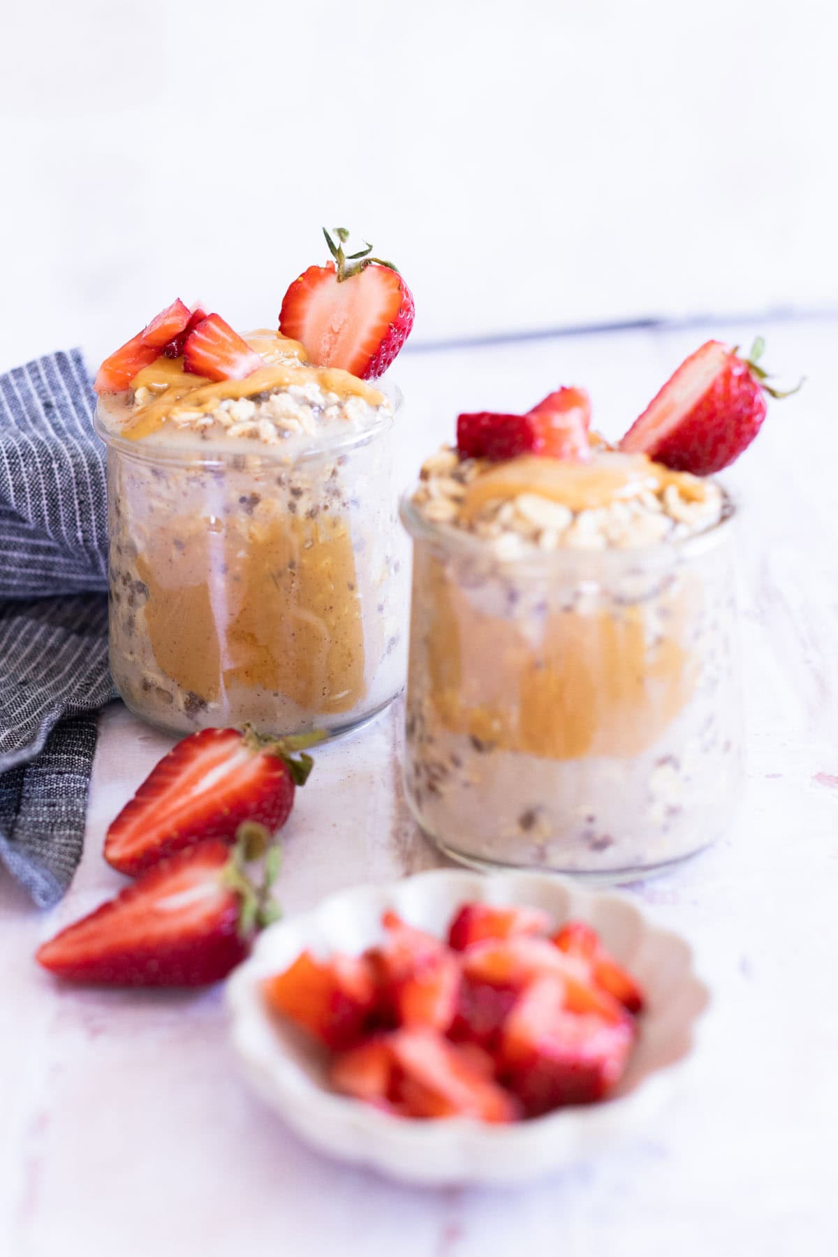 Two jars of overnight oats topped with peanut butter, chopped peanuts, and sliced strawberries, with additional strawberries and a small bowl of diced strawberries nearby.