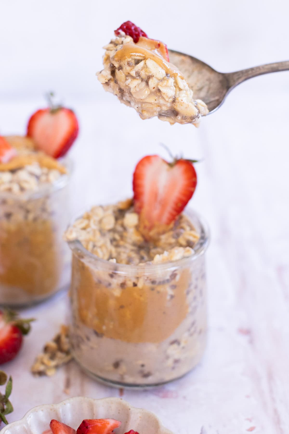 A spoonful of overnight oats with peanut butter and a strawberry slice is held above a jar filled with more overnight oats, topped with a halved strawberry.