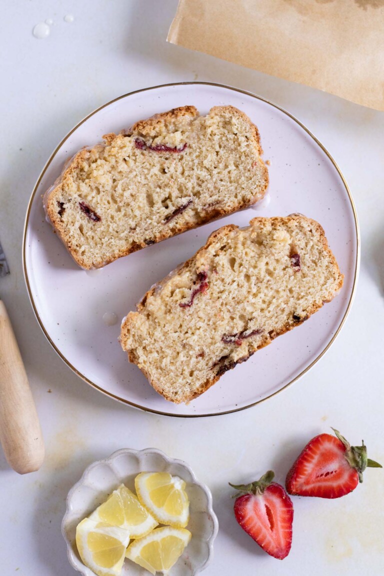 Two slices of strawberry bread are on a white plate. Next to the plate are fresh strawberries and a small dish with lemon wedges.