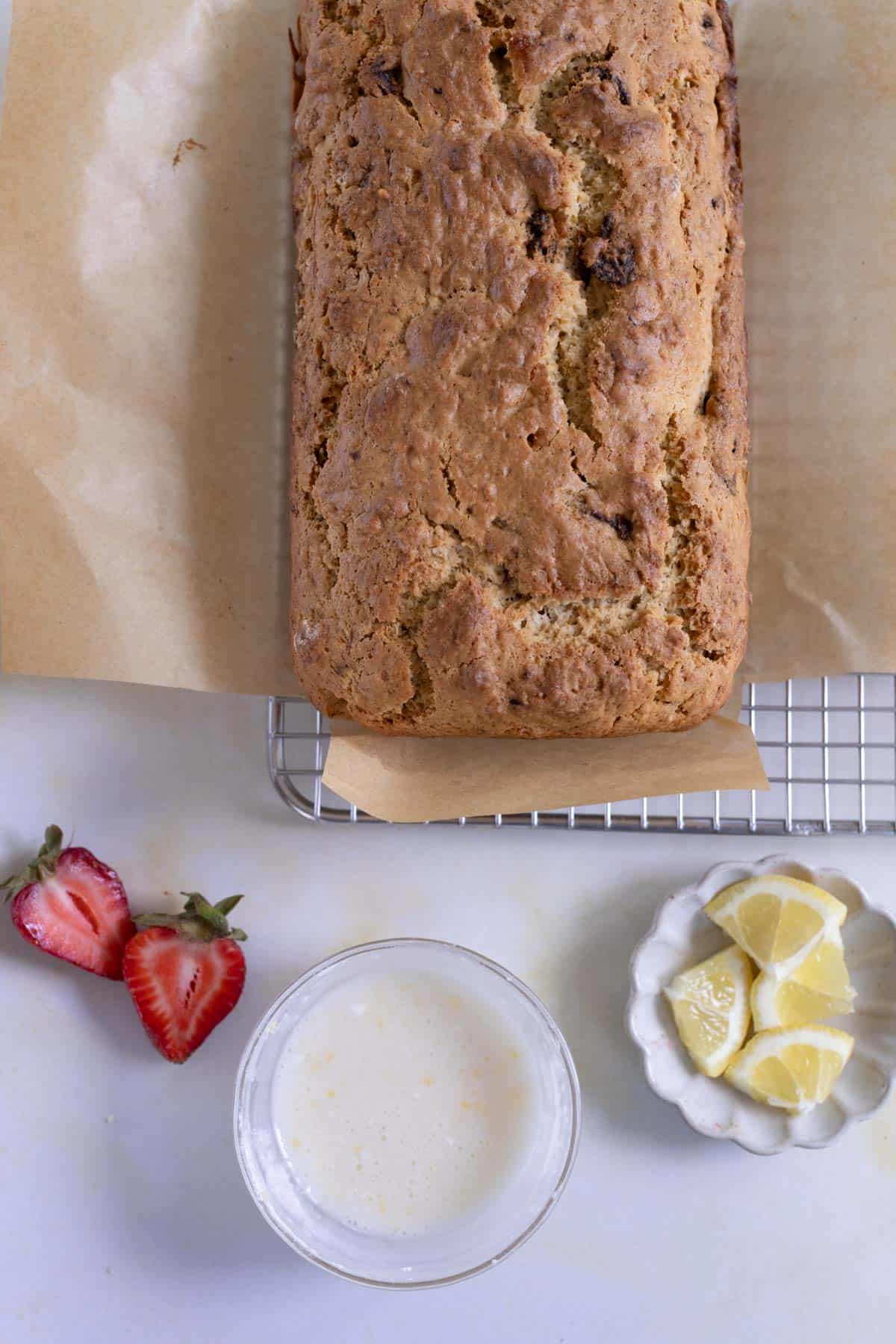 A loaf of bread on parchment paper and a cooling rack, with halved strawberries, lemon wedges in a dish, and a glass of milk on a white surface.