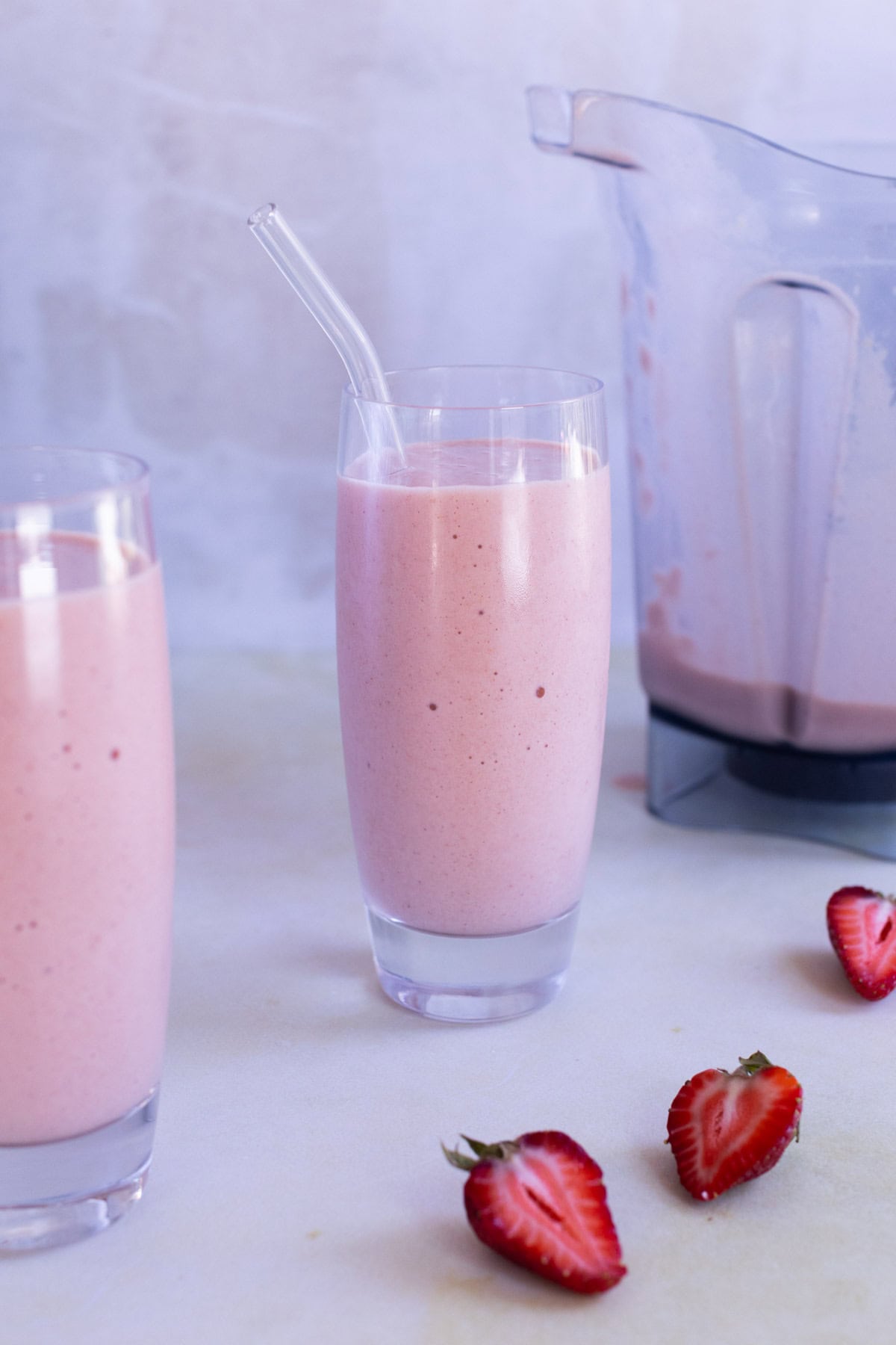 Two glasses of pink strawberry smoothie with a glass straw, a blender containing more smoothie, and sliced strawberries on a light surface.