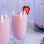 Two tall glasses of pink strawberry smoothie, one with a clear straw and a strawberry garnish, next to a blender and a small plate of sliced strawberries.