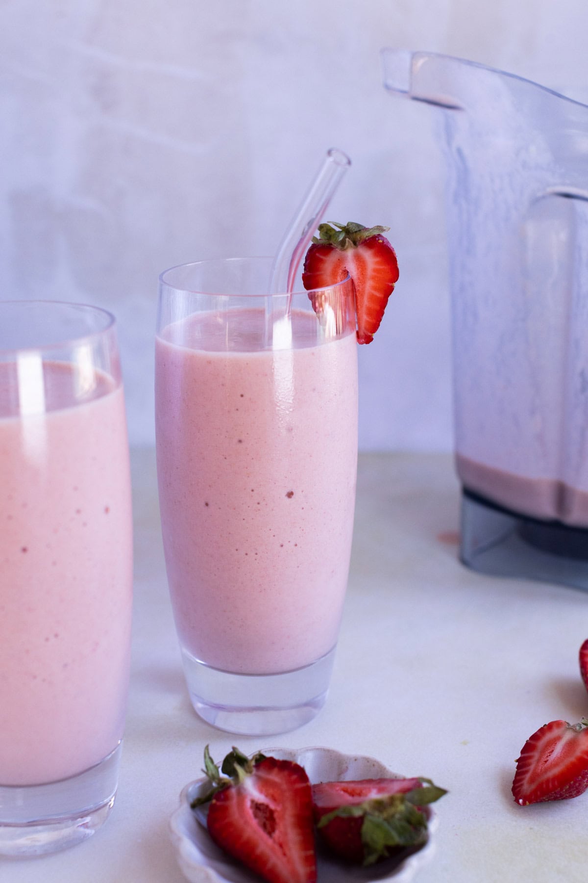 Two tall glasses of pink strawberry smoothie, one with a clear straw and a strawberry garnish, next to a blender and a small plate of sliced strawberries.