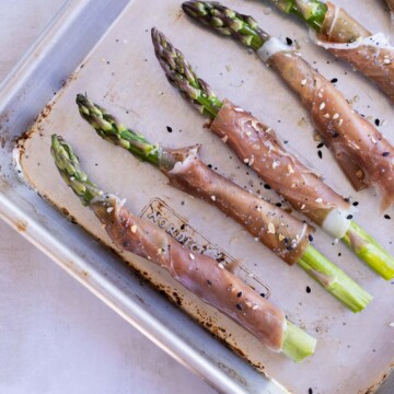 A baking tray with asparagus spears wrapped in prosciutto, sprinkled with sesame seeds, ready to be cooked.