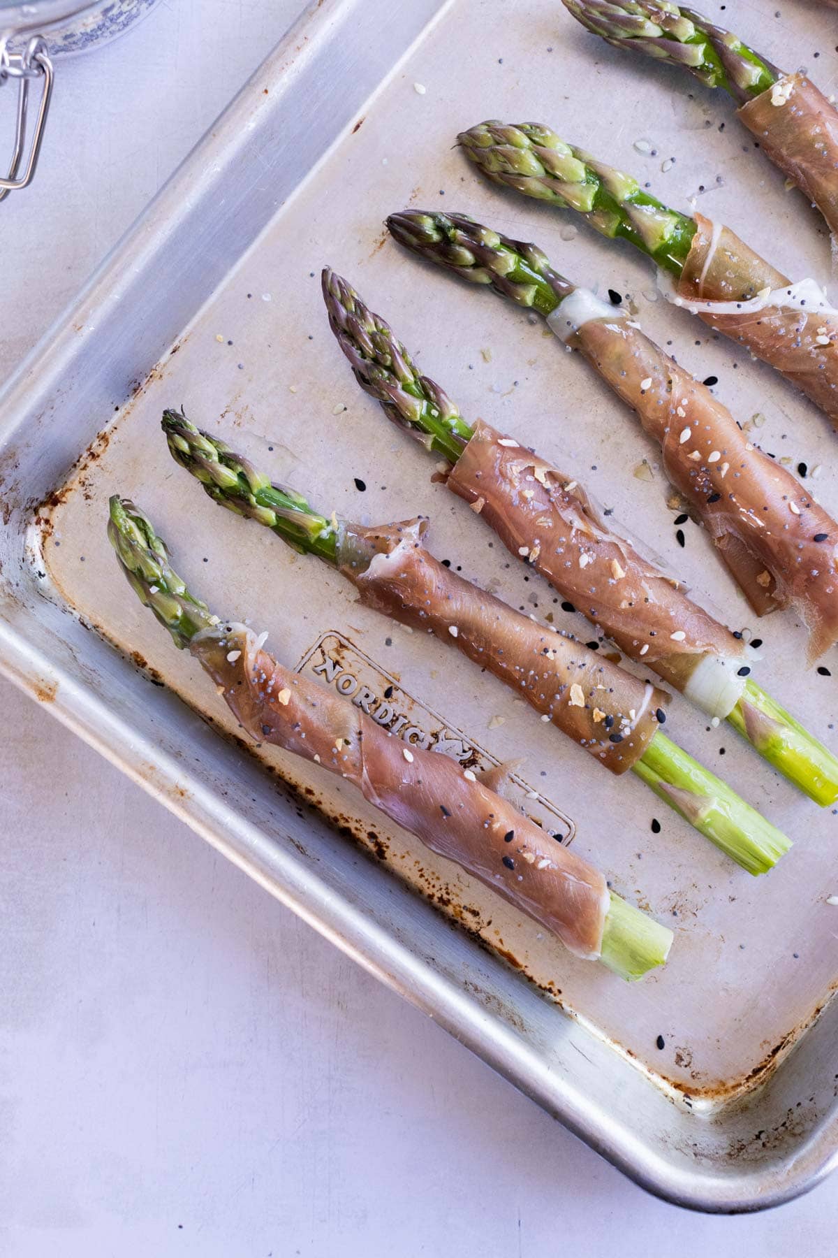 A baking tray with asparagus spears wrapped in prosciutto, sprinkled with sesame seeds, ready to be cooked.
