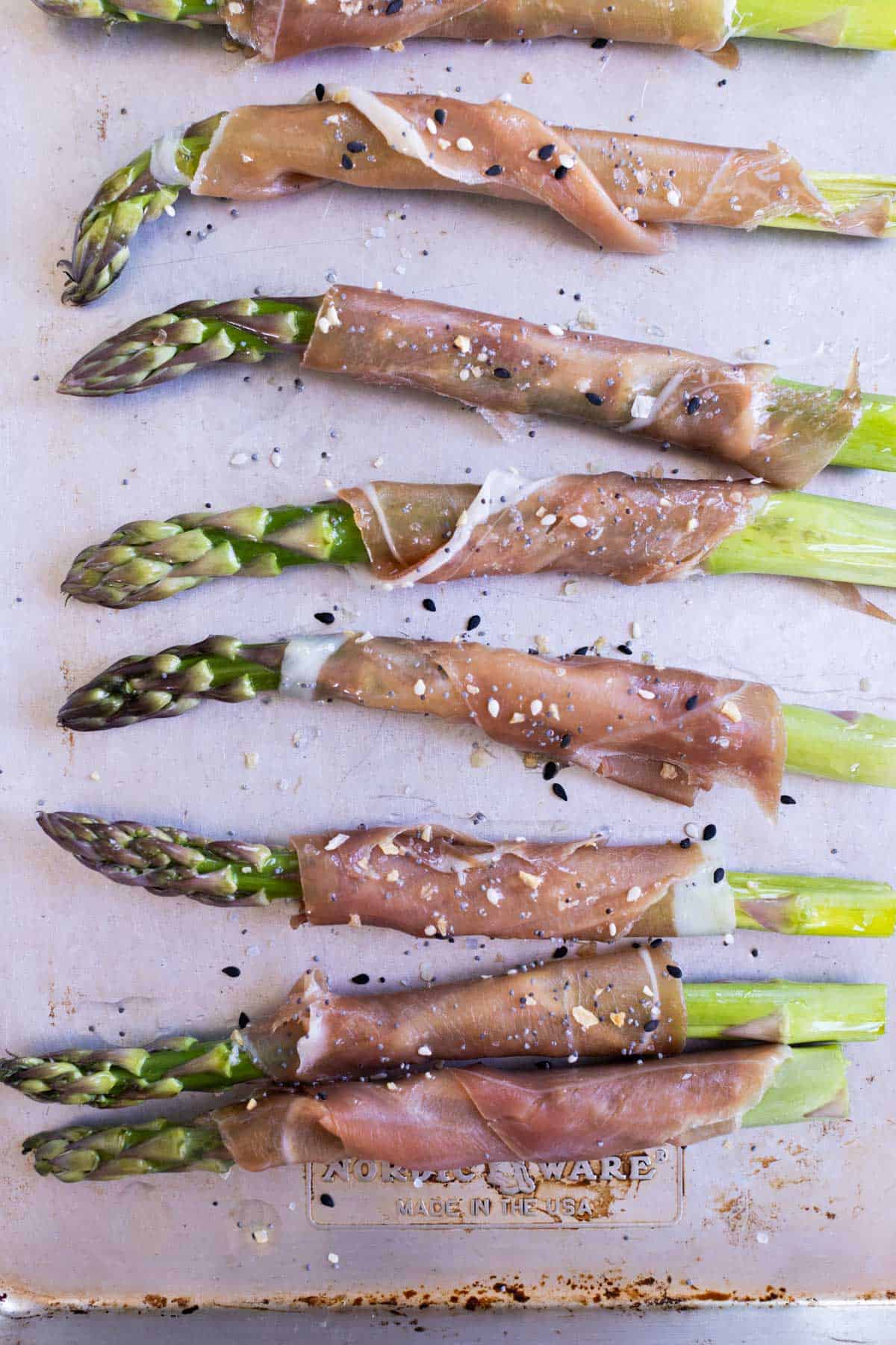 Asparagus spears wrapped in prosciutto are arranged on a metal baking sheet, sprinkled with salt, pepper, and sesame seeds.