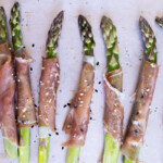 Asparagus spears wrapped in prosciutto are arranged on a baking sheet, sprinkled with sesame seeds and seasoning.