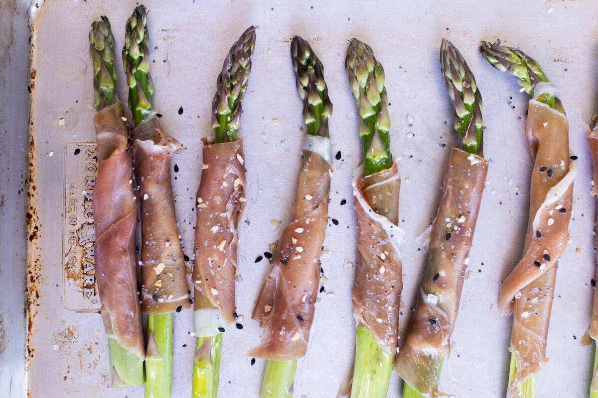 Asparagus spears wrapped in prosciutto are arranged on a baking sheet, sprinkled with sesame seeds and seasoning.