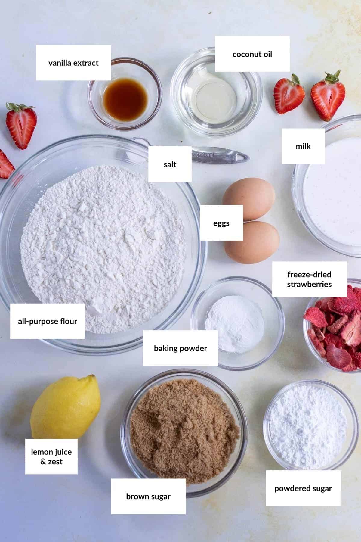Top-down view of baking ingredients in bowls, each labeled, for making strawberry bread&mdash;including flour, sugar, eggs, milk, coconut oil, strawberries, lemon, baking powder, salt, and vanilla extract.