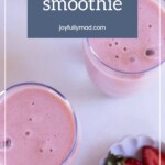 Two glasses of refreshing strawberry smoothie sit on a light surface, accompanied by a small plate with sliced strawberries.
