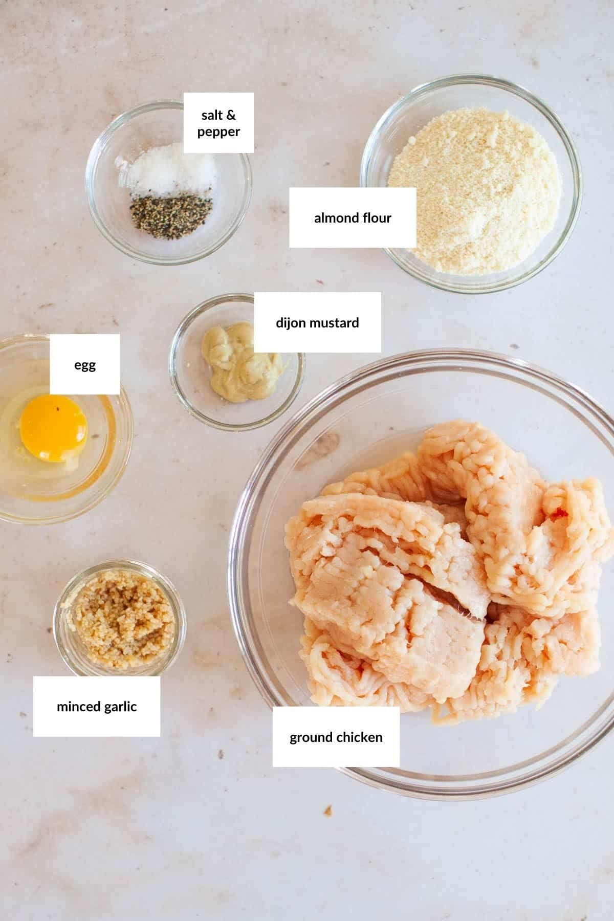 Bowls containing ground chicken, almond flour, dijon mustard, minced garlic, an egg, and salt & pepper&mdash;perfect for making chicken burgers&mdash;are arranged on a light countertop. Each ingredient is labeled.