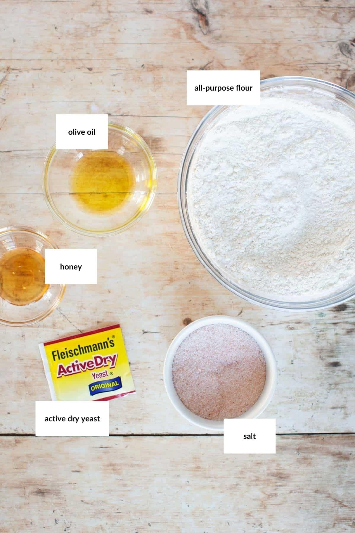 Bowls containing all-purpose flour, salt, olive oil, and honey, plus a packet of active dry yeast for thin pizza crust are arranged on a wooden surface. Each ingredient is clearly labeled.