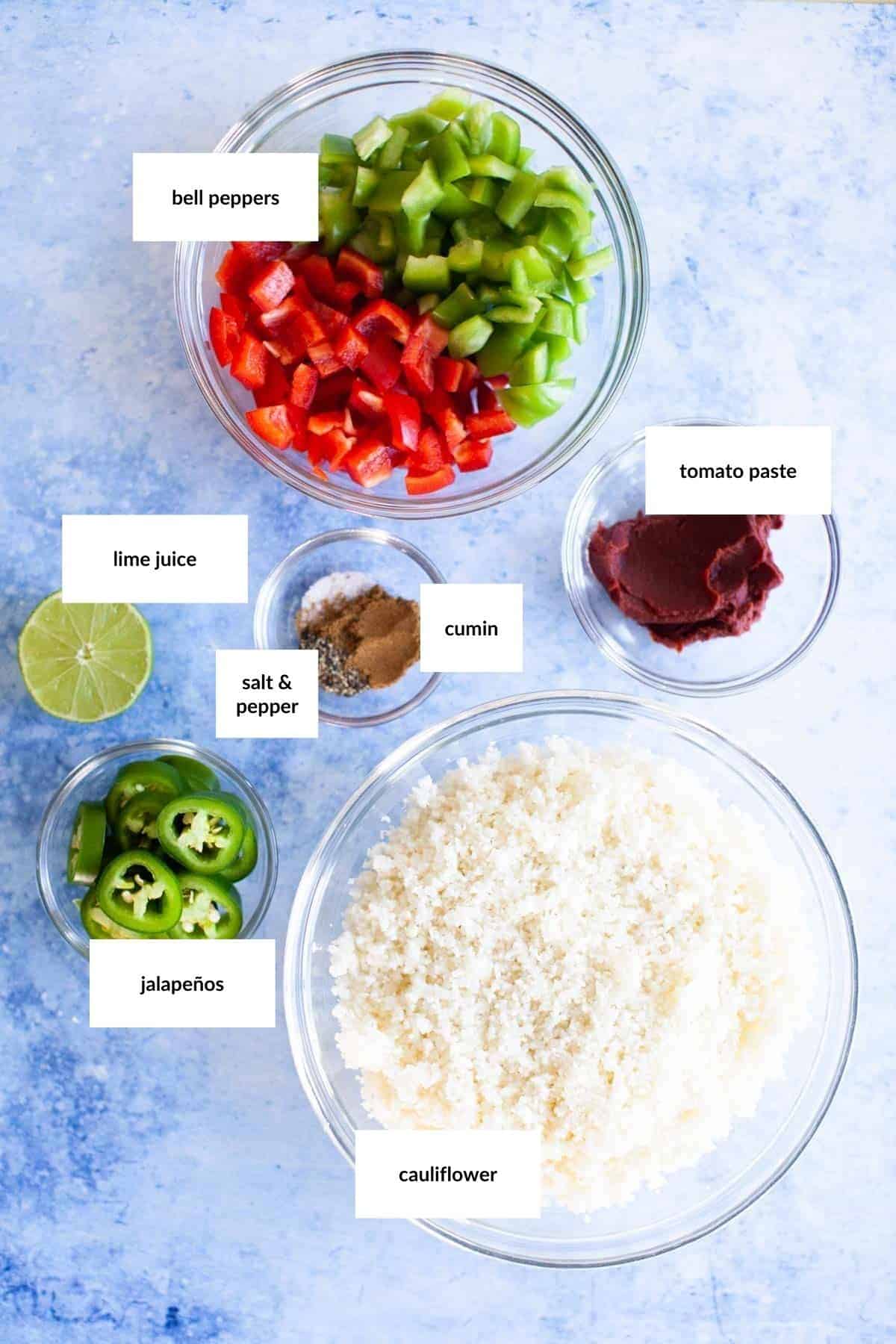 Bowls containing chopped bell peppers, tomato paste, lime juice, cumin, salt and pepper, sliced jalape&ntilde;os, and riced cauliflower on a blue background.