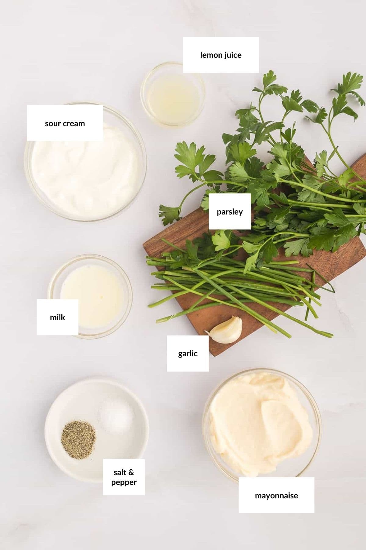Top-down view of labeled ingredients for a sauce: lemon juice, parsley, sour cream, milk, garlic, salt and pepper, and mayonnaise&mdash;perfect for dipping with a fresh vegetable tray&mdash;on a white surface.