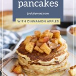 A stack of apple pancakes topped with diced cinnamon apples sits on a white plate, with a fork and a checkered cloth beside it.