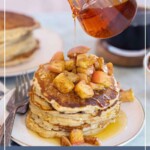 Stack of apple pancakes topped with cinnamon apples, being drizzled with syrup. More fluffy apple pancakes sit in the background, with utensils on the side.