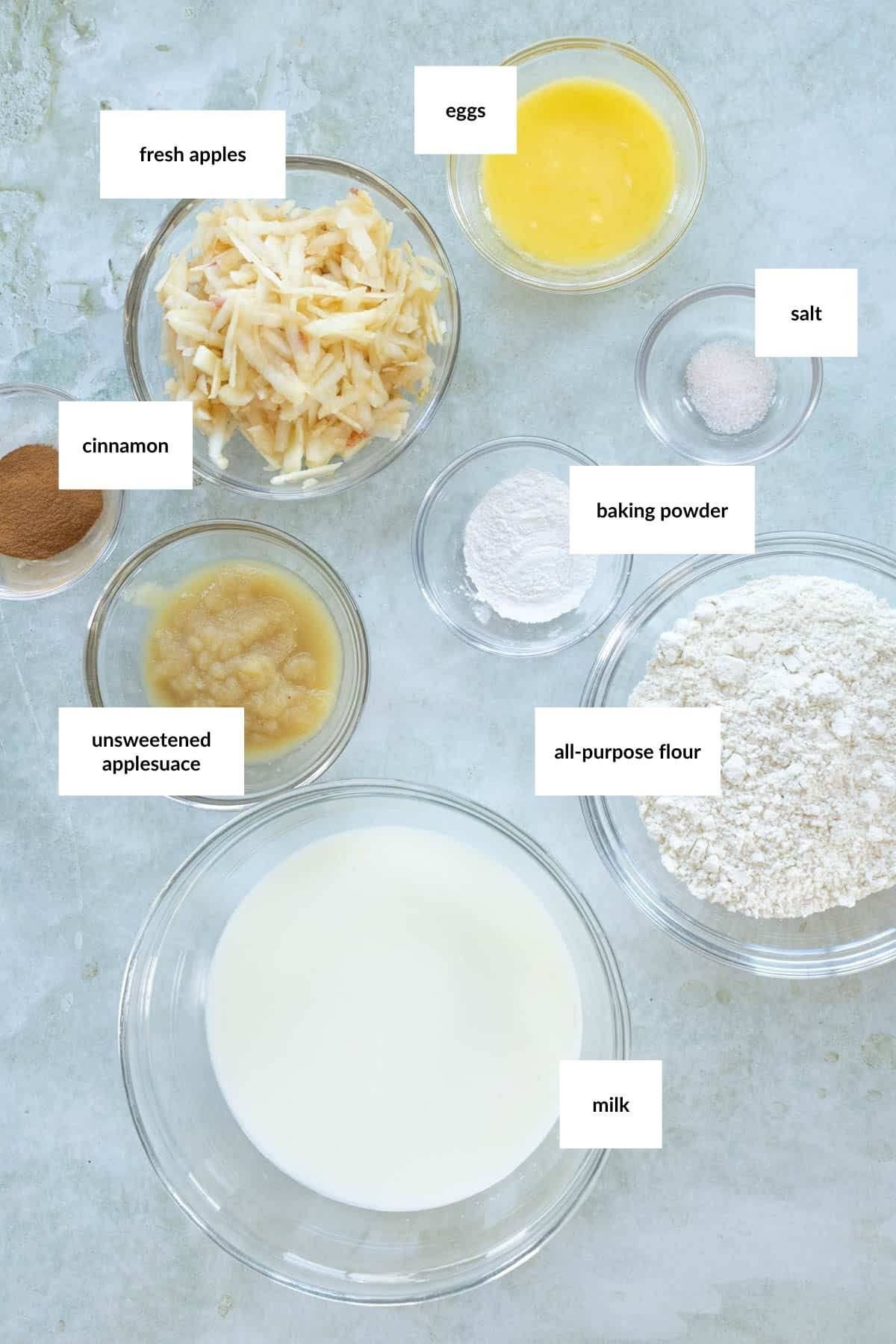 Bowls containing ingredients for apple pancakes&mdash;shredded fresh apples, beaten eggs, salt, cinnamon, baking powder, unsweetened applesauce, all-purpose flour, and milk&mdash;are arranged on a light-colored surface.
