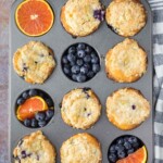 A muffin tin with nine baked blueberry muffins, three empty cups filled with fresh blueberries and orange slices, on a textured surface with a striped towel nearby.