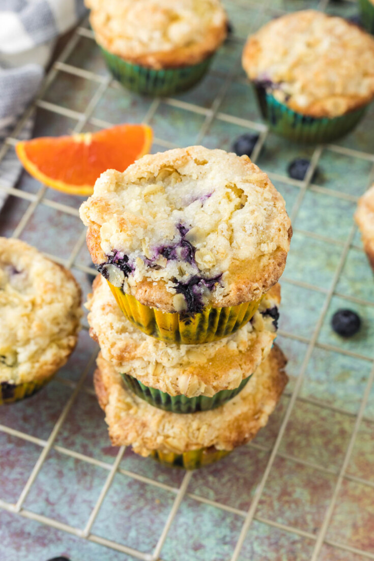 Three blueberry orange muffins with crumb topping stacked on a cooling rack, with loose blueberries and a slice of orange nearby.