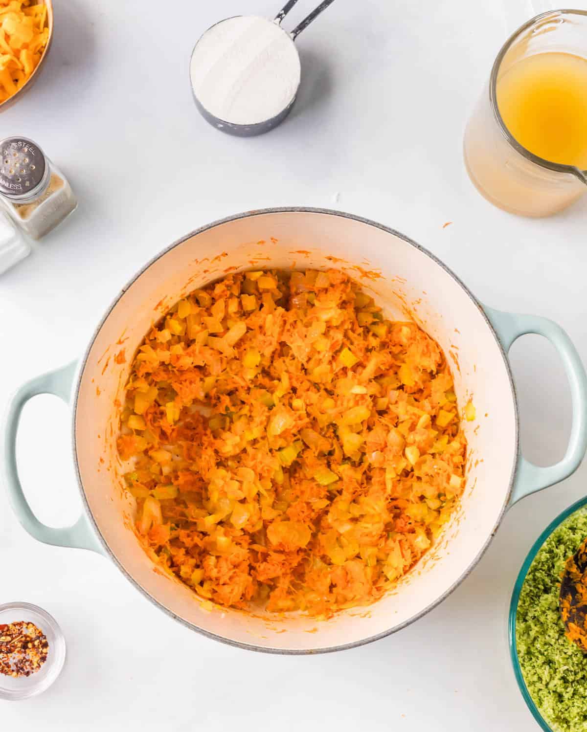 A pot filled with saut&eacute;ed onions and carrots, surrounded by a measuring cup of broth, a bowl of chopped greens, a cup of flour, and small bowls of seasonings on a white surface.