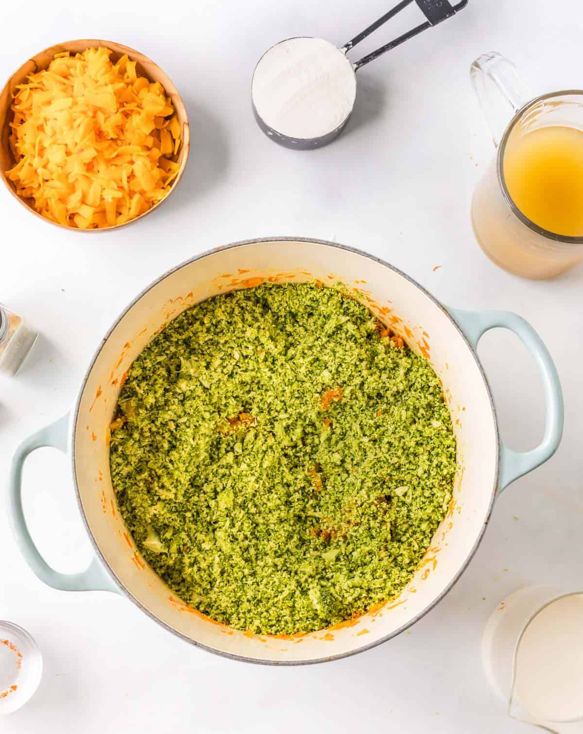 A pot filled with finely chopped broccoli and carrots on a white surface, surrounded by bowls of shredded cheese, measuring cup, and a pitcher of broth.