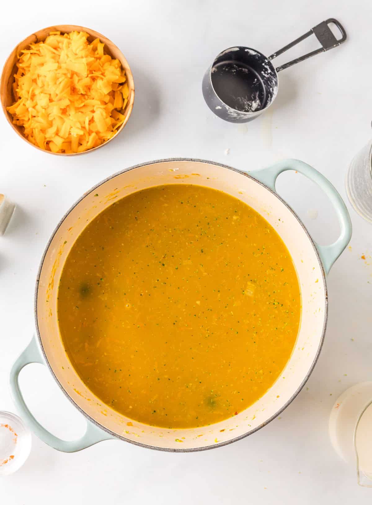 A pot of yellow soup sits on a white surface, surrounded by a bowl of shredded cheese, a measuring cup, and other cooking utensils.