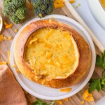 A bread bowl filled with broccoli cheddar soup, topped with shredded cheddar cheese, on a wooden board with broccoli and parsley nearby.