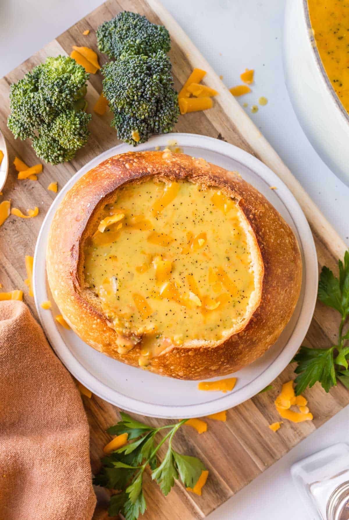 A bread bowl filled with broccoli cheddar soup, topped with shredded cheddar cheese, on a wooden board with broccoli and parsley nearby.