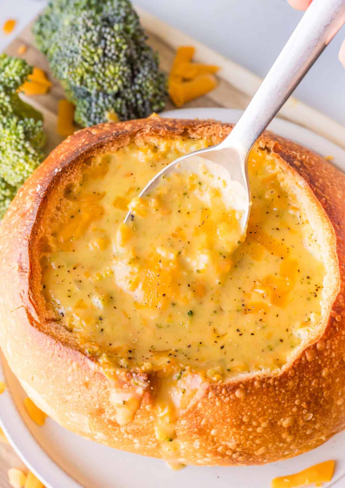 A spoon scooping creamy broccoli cheddar soup from a bread bowl, with broccoli florets and shredded cheese on the side.