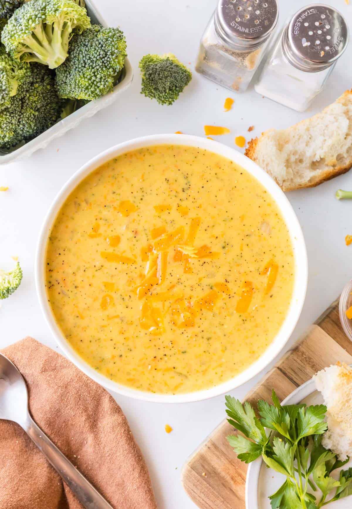 A bowl of broccoli cheddar soup garnished with shredded cheese is on a white table, surrounded by broccoli, bread, parsley, and salt and pepper shakers.