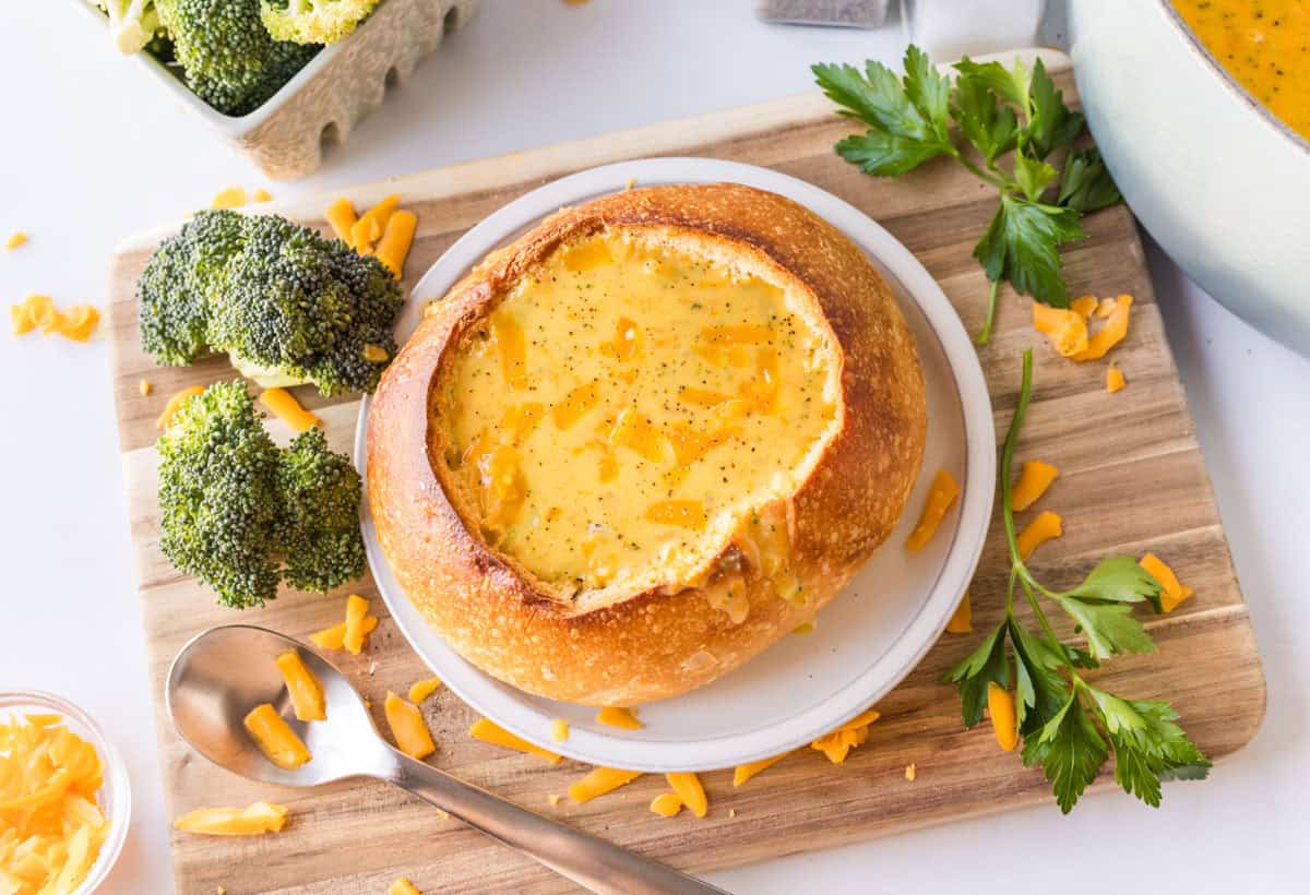 A bread bowl filled with broccoli cheddar soup sits on a plate, surrounded by fresh broccoli, shredded cheddar cheese, and parsley on a wooden board.